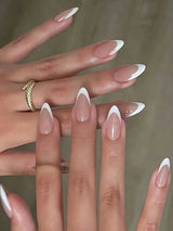 Close-up of hands with long almond-shaped press-on nails in a classic French tip style, featuring a glossy nude-pink base and crisp white tips. One hand wears a textured gold ring against a soft beige background.