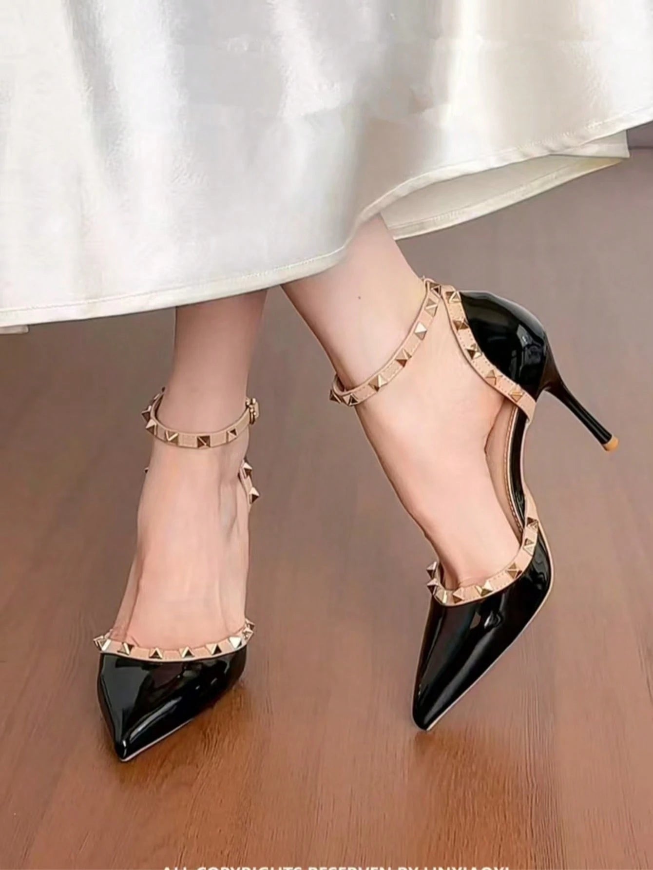 Black pointed-toe stiletto heels with gold-tone ankle straps and pyramid studs, shown on a model wearing a white skirt over a wooden floor.