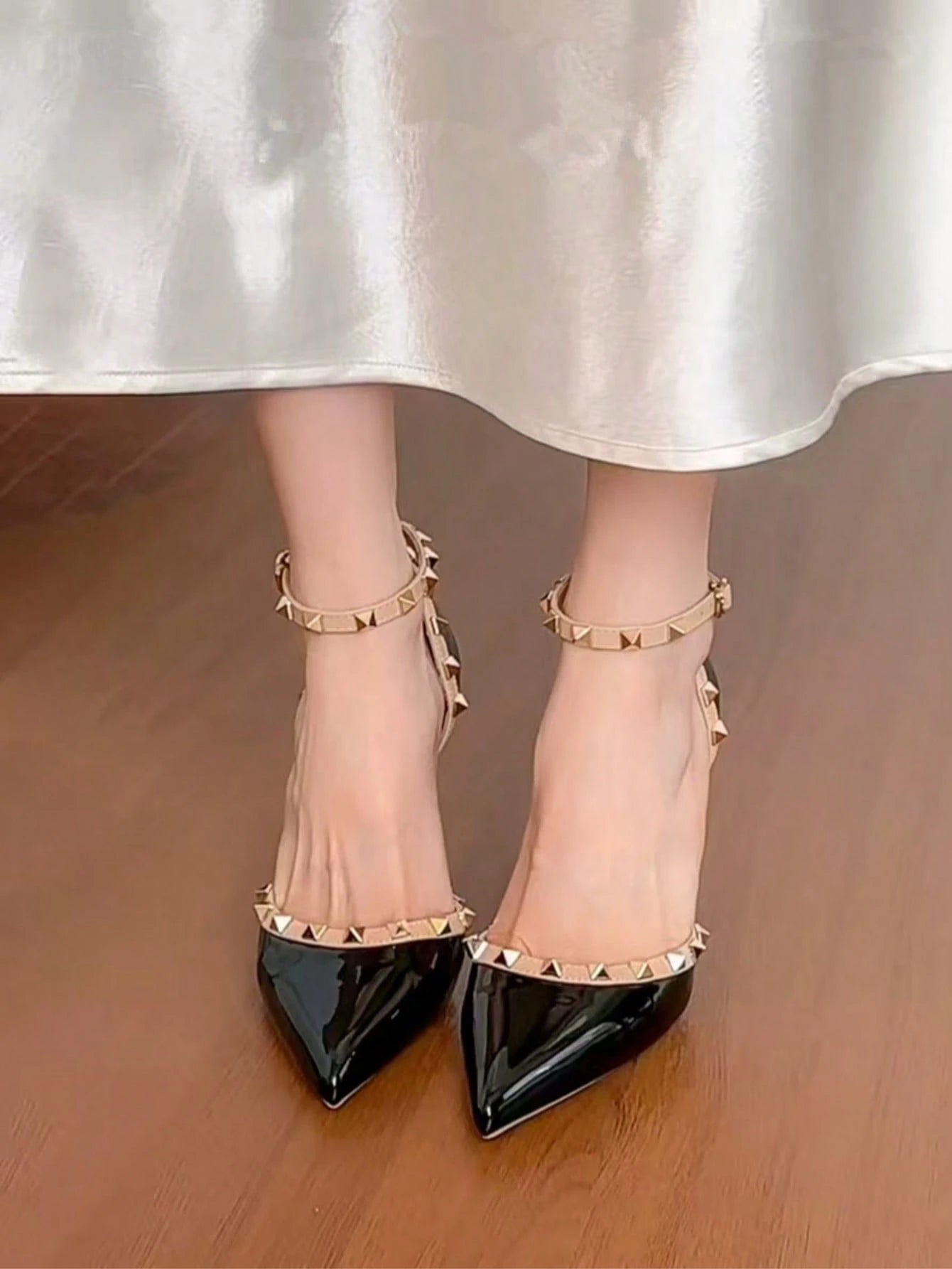 Black pointed-toe shoes with gold studded straps worn with a white satin skirt.