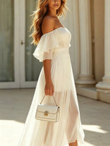 A woman wearing a flowing off-the-shoulder white maxi dress with a ruffled sleeve, holding a white structured handbag with gold hardware, set against a sunlit classical building background with columns.