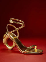 A single metallic gold high-heeled sandal with an abstract sculptural heel and thin wraparound ankle straps, photographed on a dark red studio background.