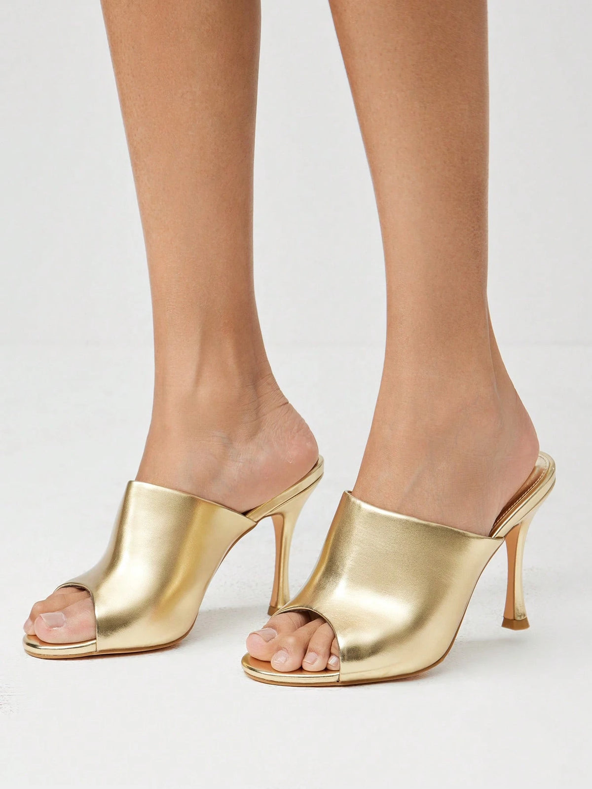 A pair of metallic gold open-toe mule high heels shown on a model's feet against a light gray studio background.