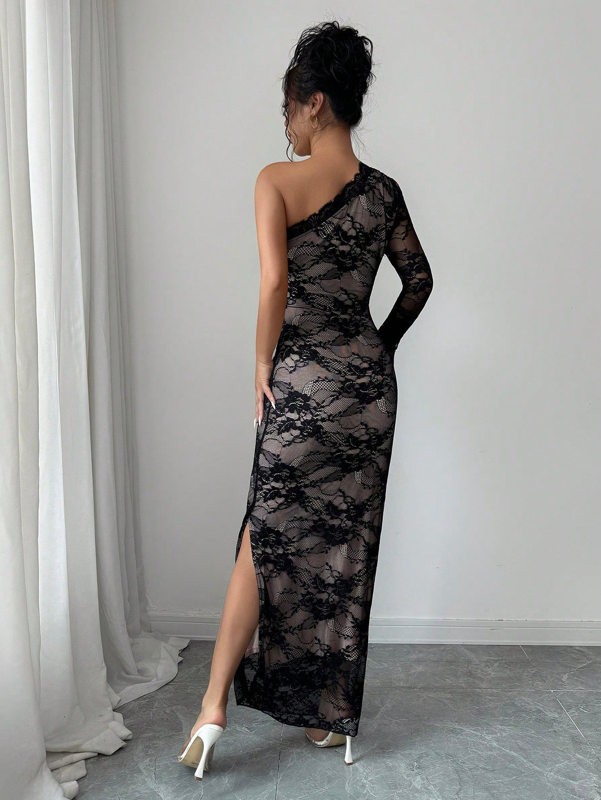 A woman wearing a black lace one-shoulder maxi dress with a thigh-high slit, shown from the back in a room with a gray floor and white curtains.