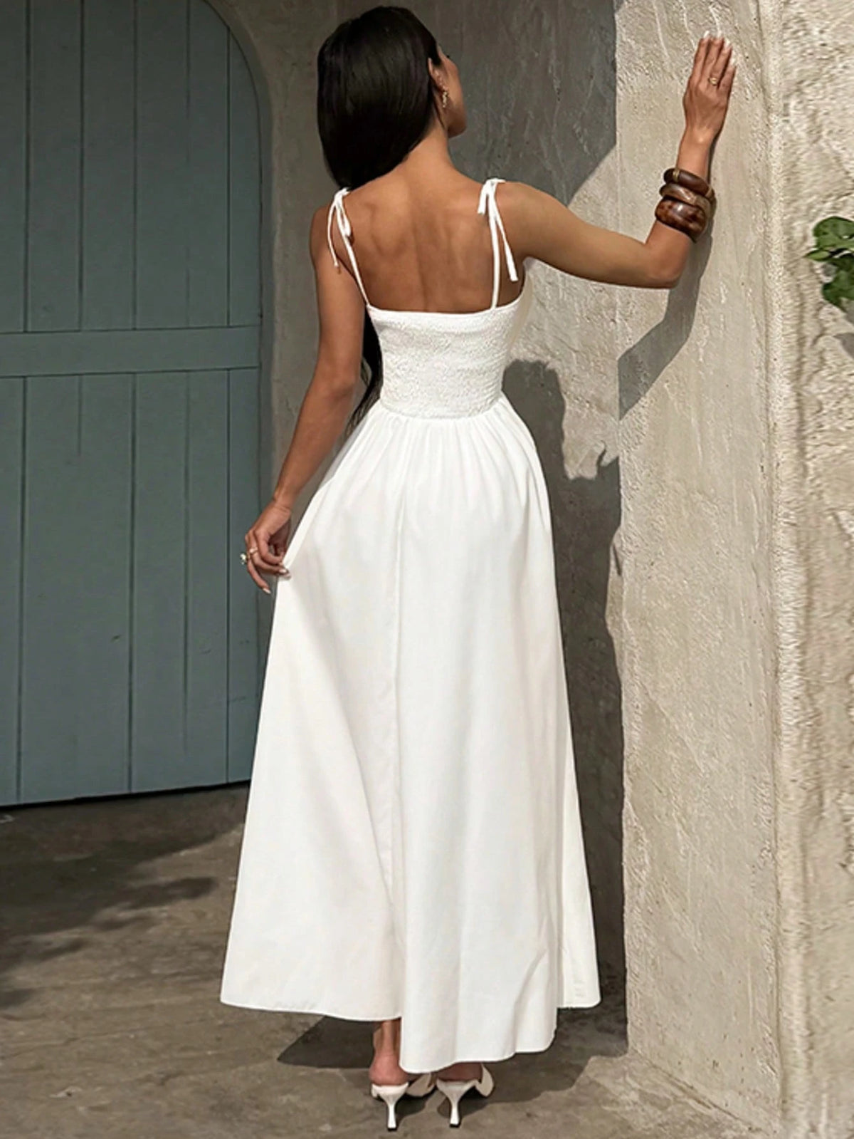 A woman wearing a white sleeveless maxi dress with thin shoulder straps and a fitted waist, photographed from the back in an outdoor setting against a textured wall.