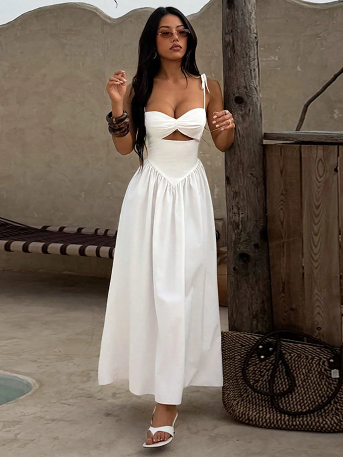 A woman models a white sleeveless maxi dress with a sweetheart neckline and a midriff cutout, standing in a rustic outdoor setting with a woven bag and wooden post.