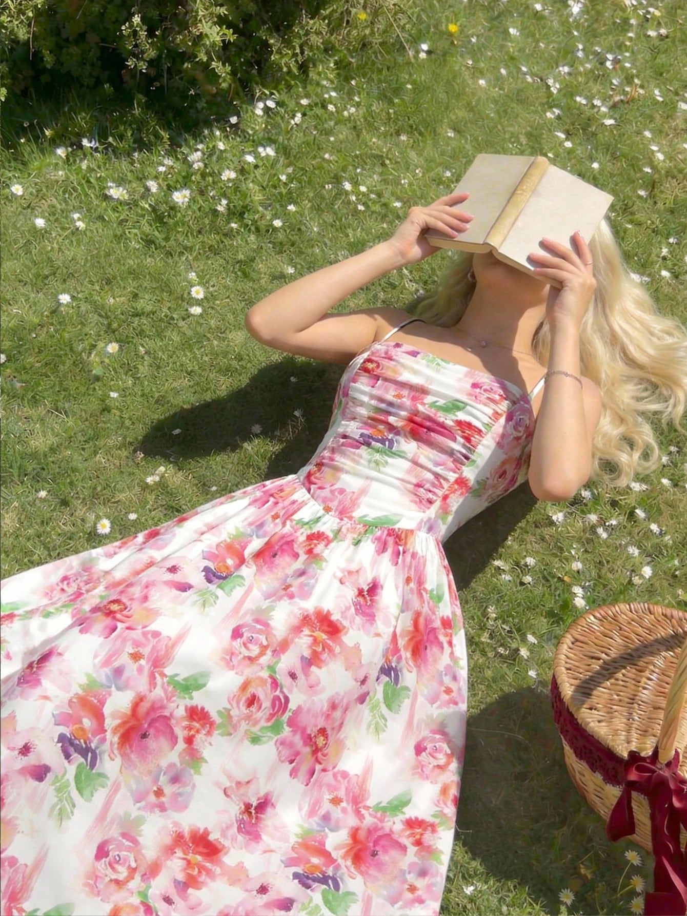 Woman in a floral dress reading a book outdoors on grass with flowers.