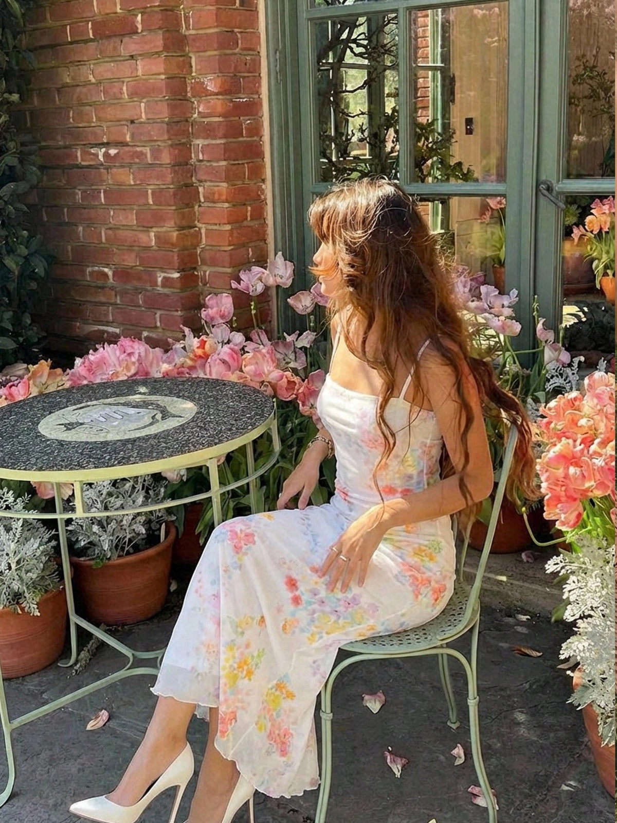 A woman sits on a light green metal patio chair next to a small round table in an outdoor garden setting. She is wearing a white strapless midi dress with a pastel pink, yellow, and blue floral print, paired with white pointed-toe heels.