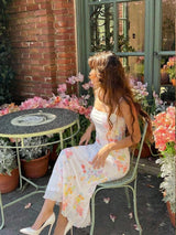 A woman sits on a light green metal patio chair next to a small round table in an outdoor garden setting. She is wearing a white strapless midi dress with a pastel pink, yellow, and blue floral print, paired with white pointed-toe heels.