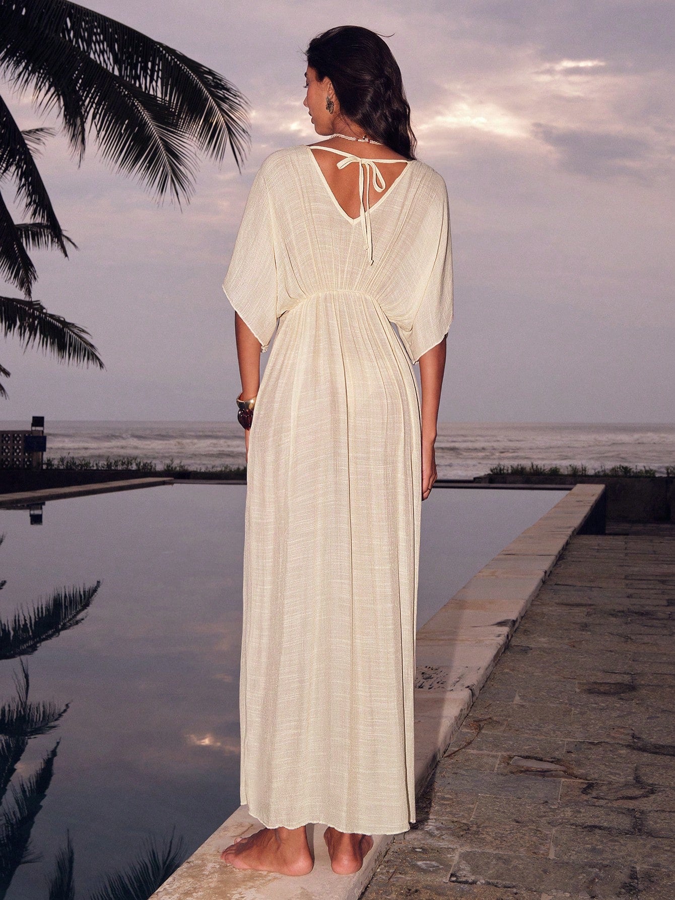 Woman in a long white dress standing by a pool with palm trees and ocean in the background