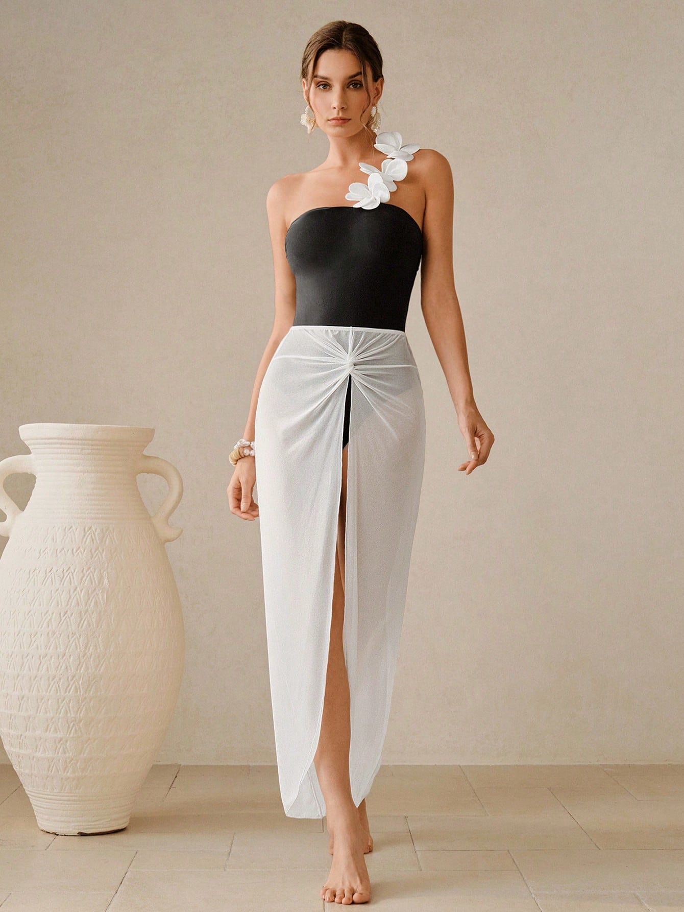 Woman wearing a black top and white skirt with a flower in her hair, standing next to a large white vase.