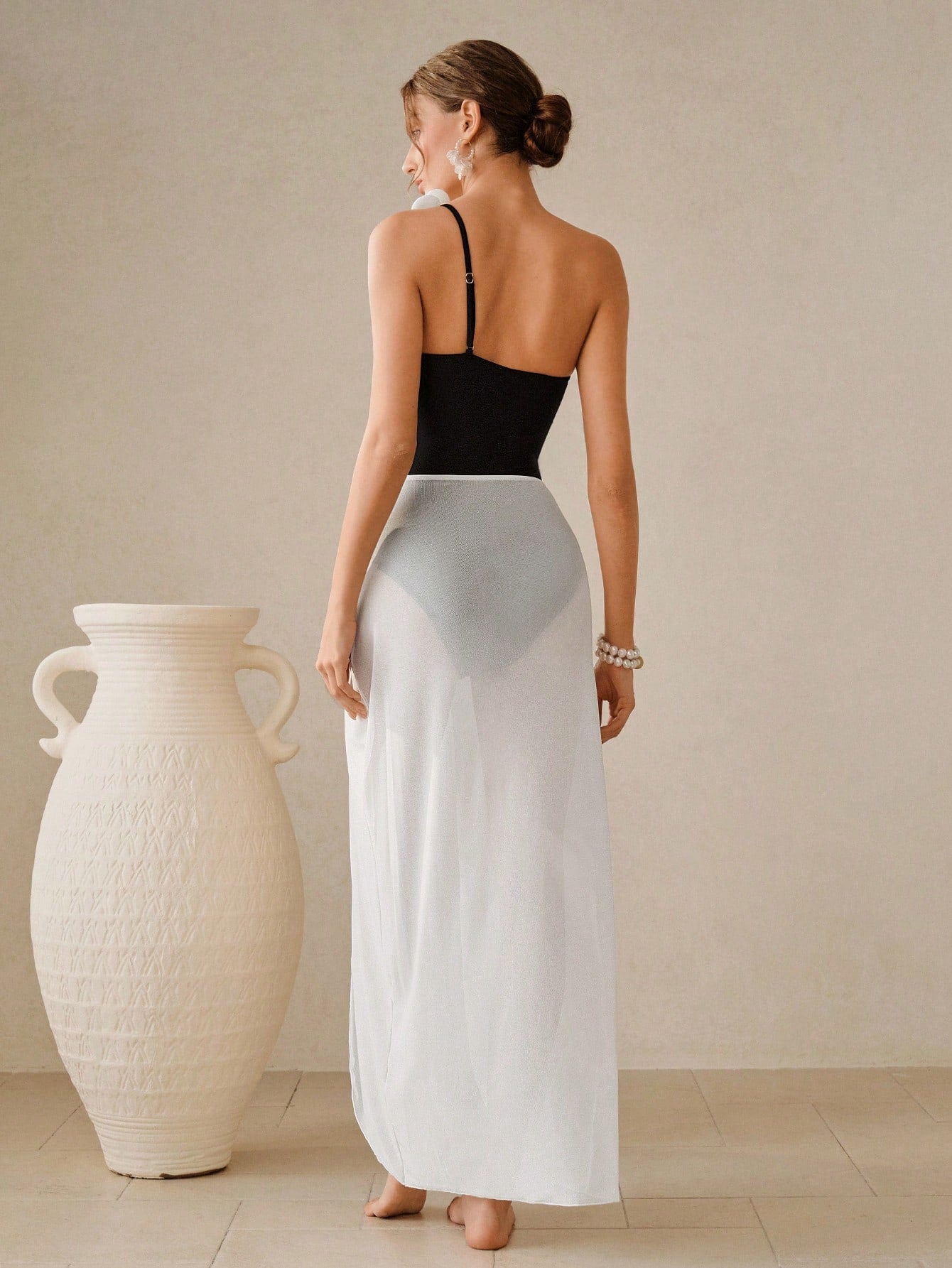 Woman in a black top and white skirt standing next to a large white vase against a beige wall.