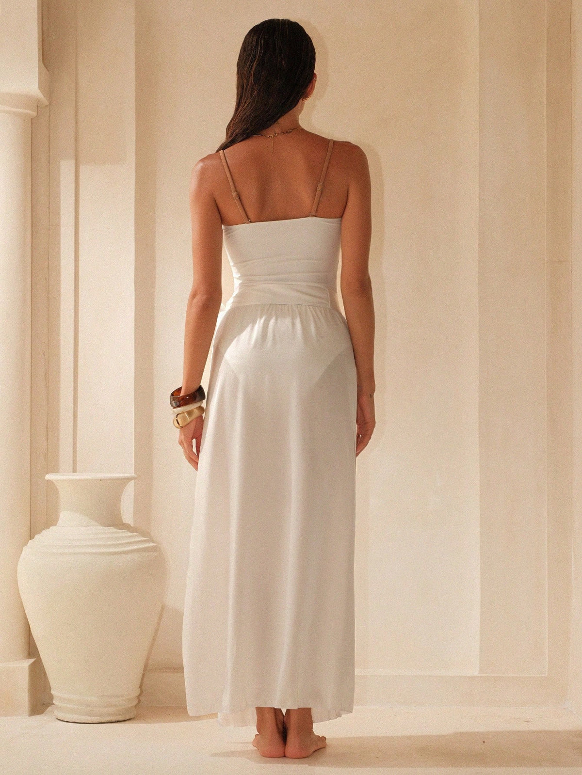 A woman stands with her back to the camera wearing a white spaghetti-strap maxi dress in a warm, sunlit interior with a large white vase on the left.