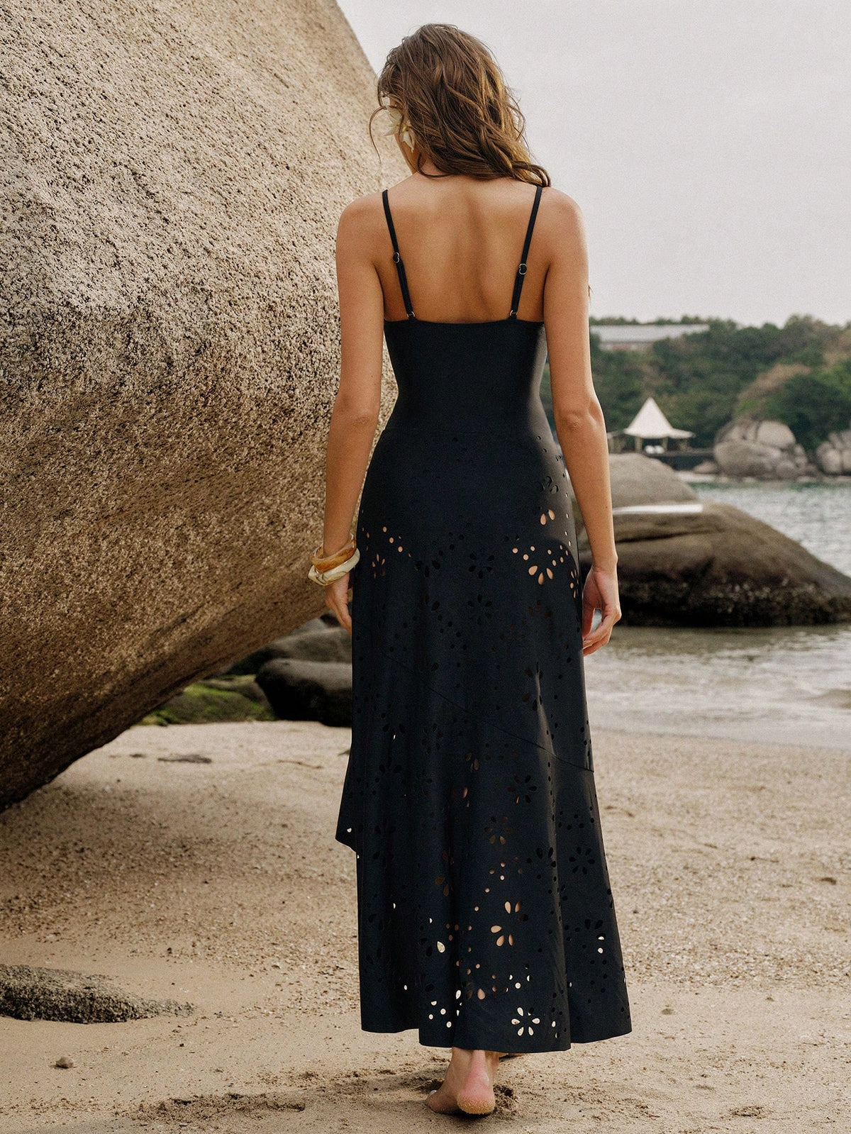 A woman stands on a sandy beach with large rocks and water in the background, wearing a black sleeveless maxi dress with cutout details and thin straps, shown from the back.