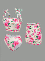 A three-piece bikini set featuring a pink and green floral print on a white base, including a tie-strap crop top, high-waisted mini skirt, and high-waisted bottoms, displayed against a solid gray background.