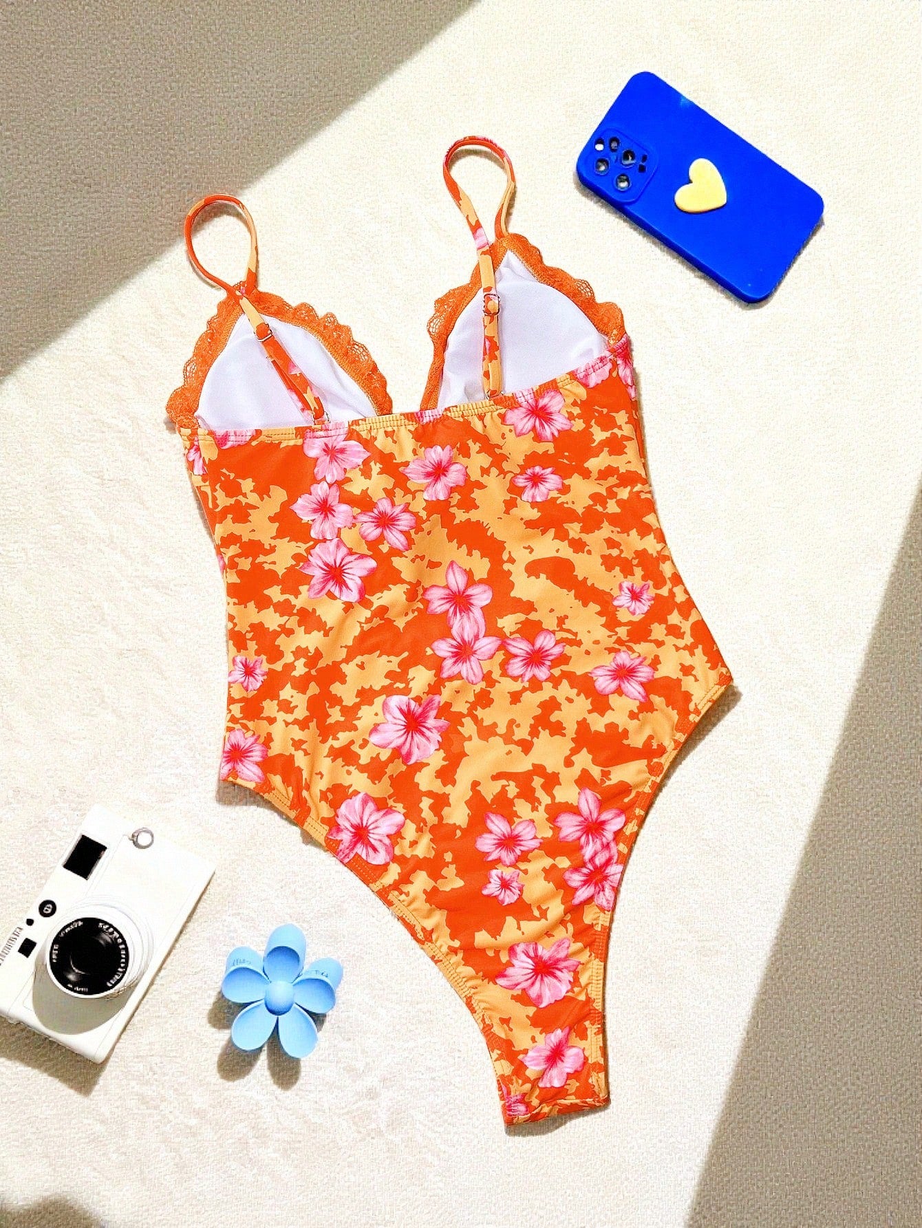 Orange floral swimsuit on a white surface with a blue phone and camera.