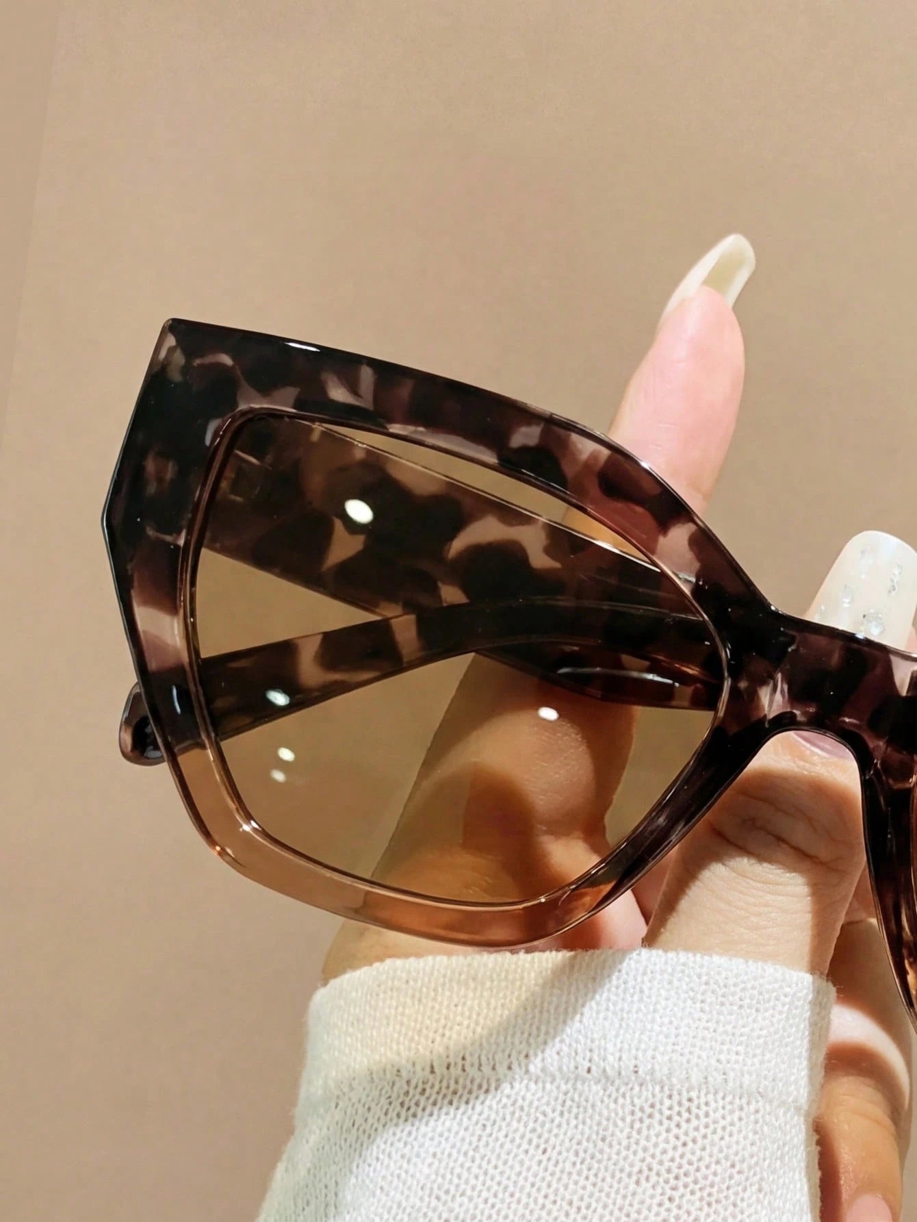 Sunglasses with tortoiseshell frame and brown lenses held by a hand against a beige background