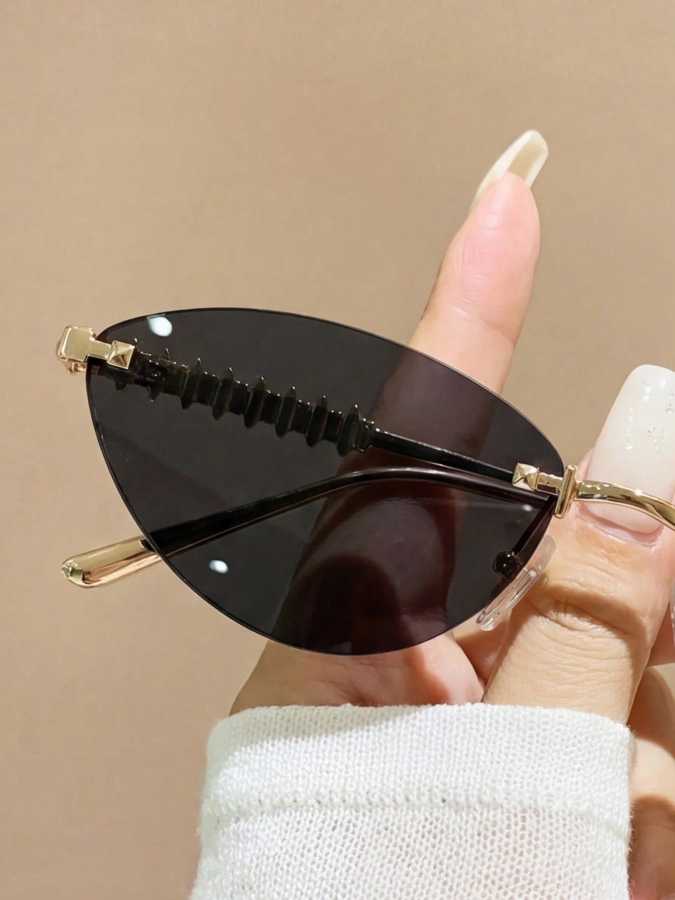 Sunglasses with black lenses and gold frames held by a hand against a beige background