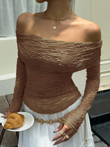 A woman wearing a fitted off-the-shoulder long-sleeve top in a warm taupe-brown crinkled fabric, paired with a white skirt and gold jewelry, holding a croissant.