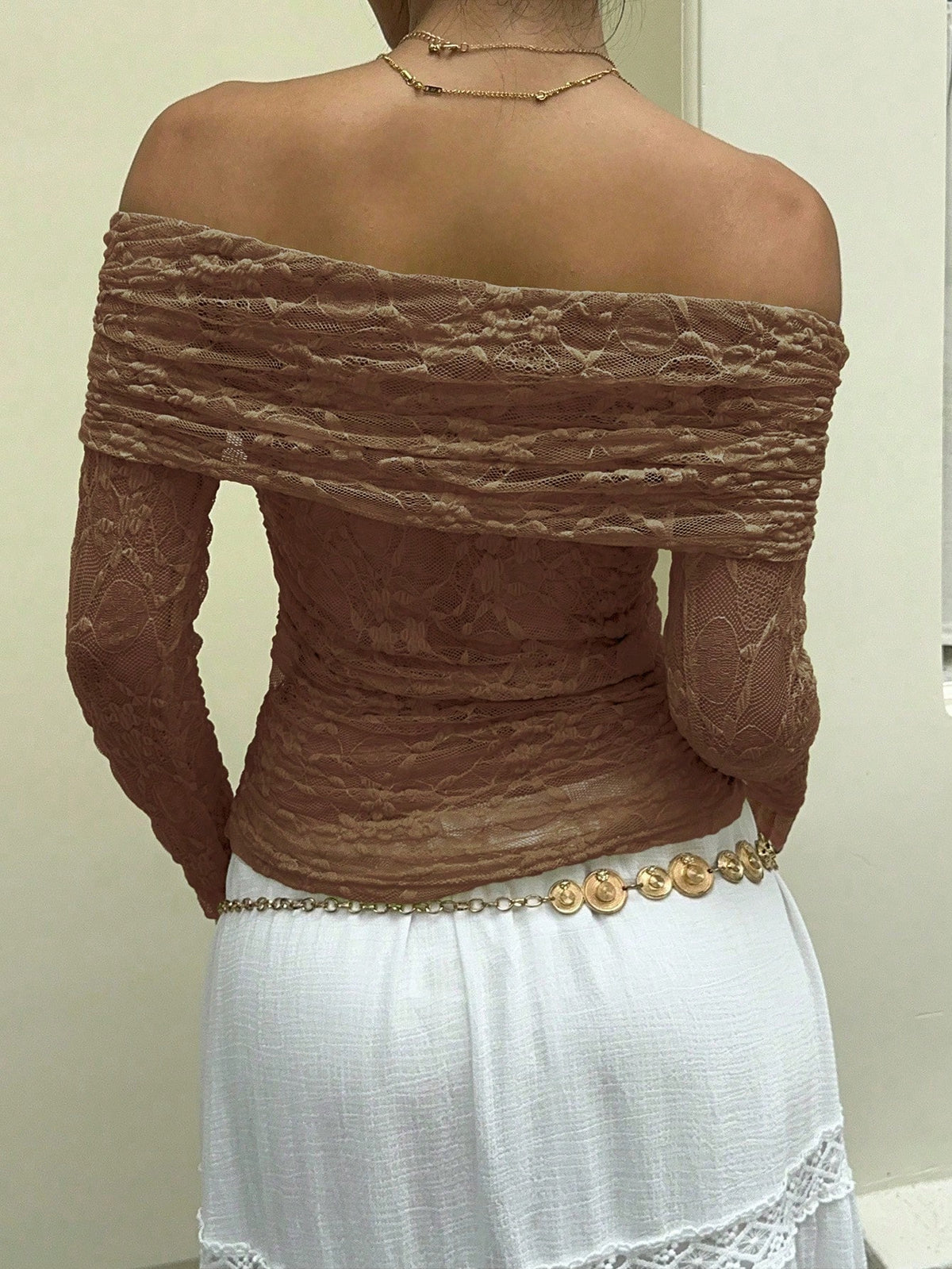 A woman shown from the back wearing a brown off-the-shoulder lace long-sleeve top and a white skirt with a gold coin belt.