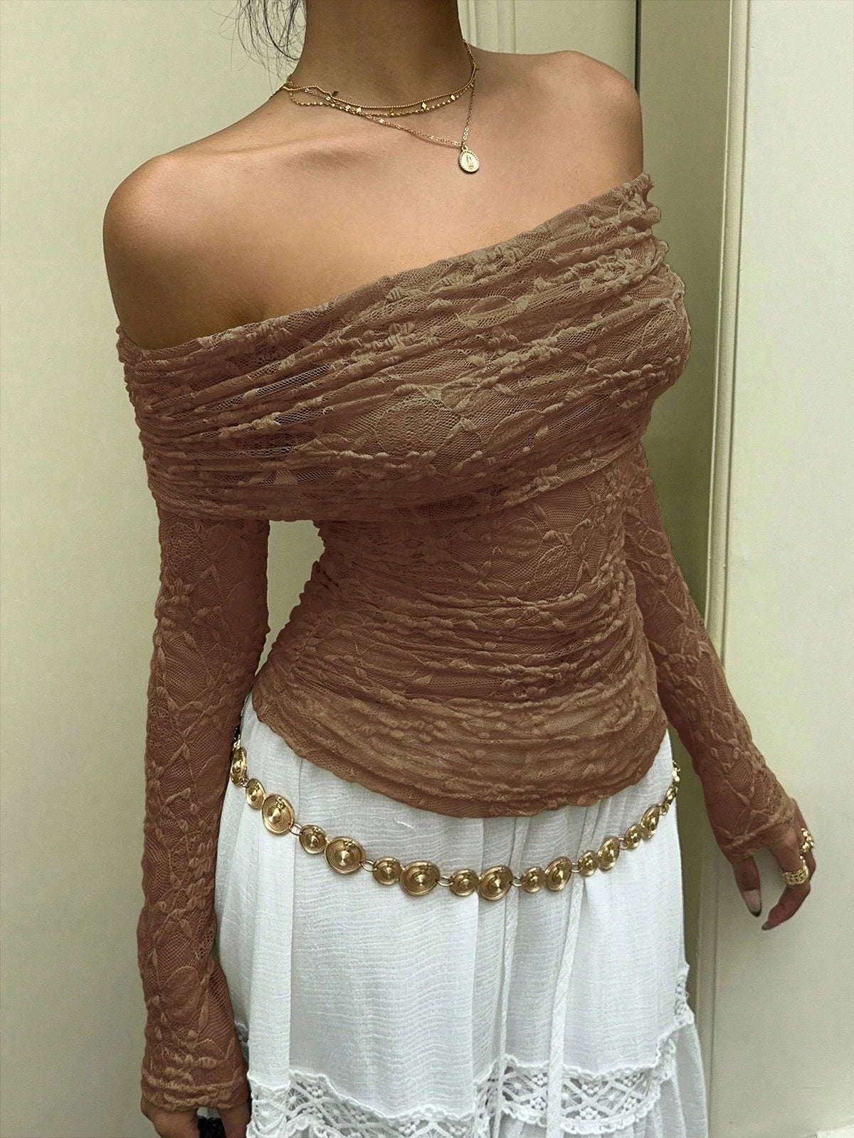 A woman wearing a fitted off-the-shoulder long-sleeve top in a warm taupe-brown lace-like fabric, paired with a white skirt and a gold beaded belt.
