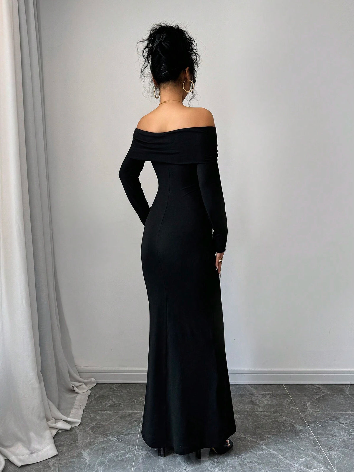 A woman stands with her back to the camera wearing a long black off-the-shoulder maxi dress in a minimalist room with a gray tiled floor and white walls.