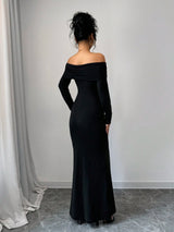 A woman stands with her back to the camera wearing a long black off-the-shoulder maxi dress in a minimalist room with a gray tiled floor and white walls.