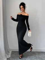 A woman modeling a long black off-the-shoulder maxi dress with a large floral detail at the neckline, holding a small white feathered clutch in a minimalist indoor setting.