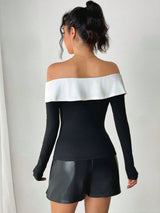 A woman shown from the back wearing a black long-sleeve off-the-shoulder top with a wide white band across the shoulders, paired with black shorts, against a plain light gray wall.