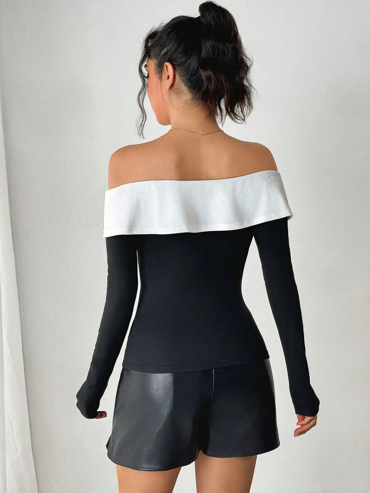 A woman shown from the back wearing a black long-sleeve off-the-shoulder top with a wide white band across the shoulders, paired with black shorts, against a plain light gray wall.