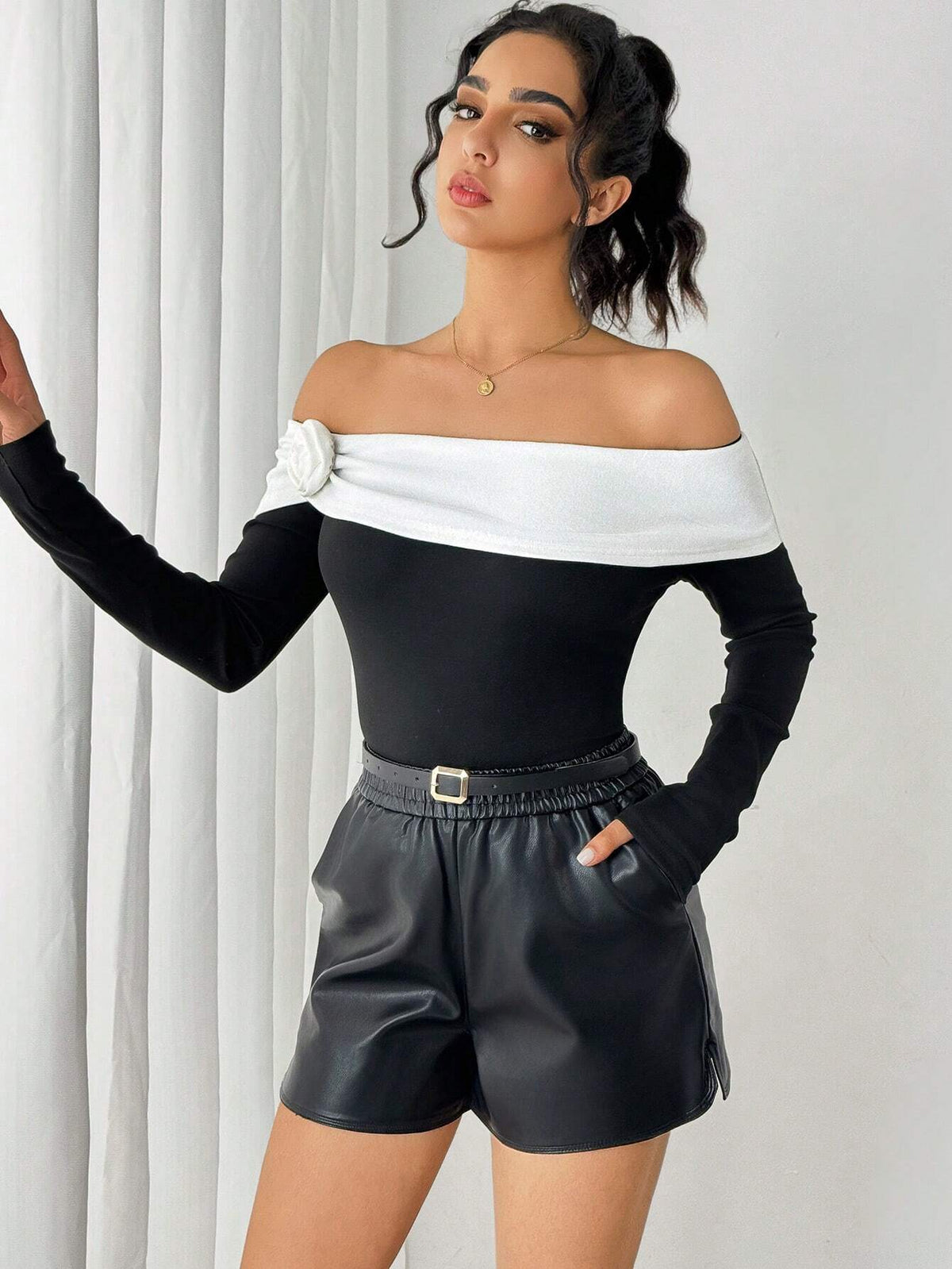 A woman modeling an off-the-shoulder black long-sleeve top with a wide white band across the chest, paired with black faux-leather shorts, posed in front of white curtains.