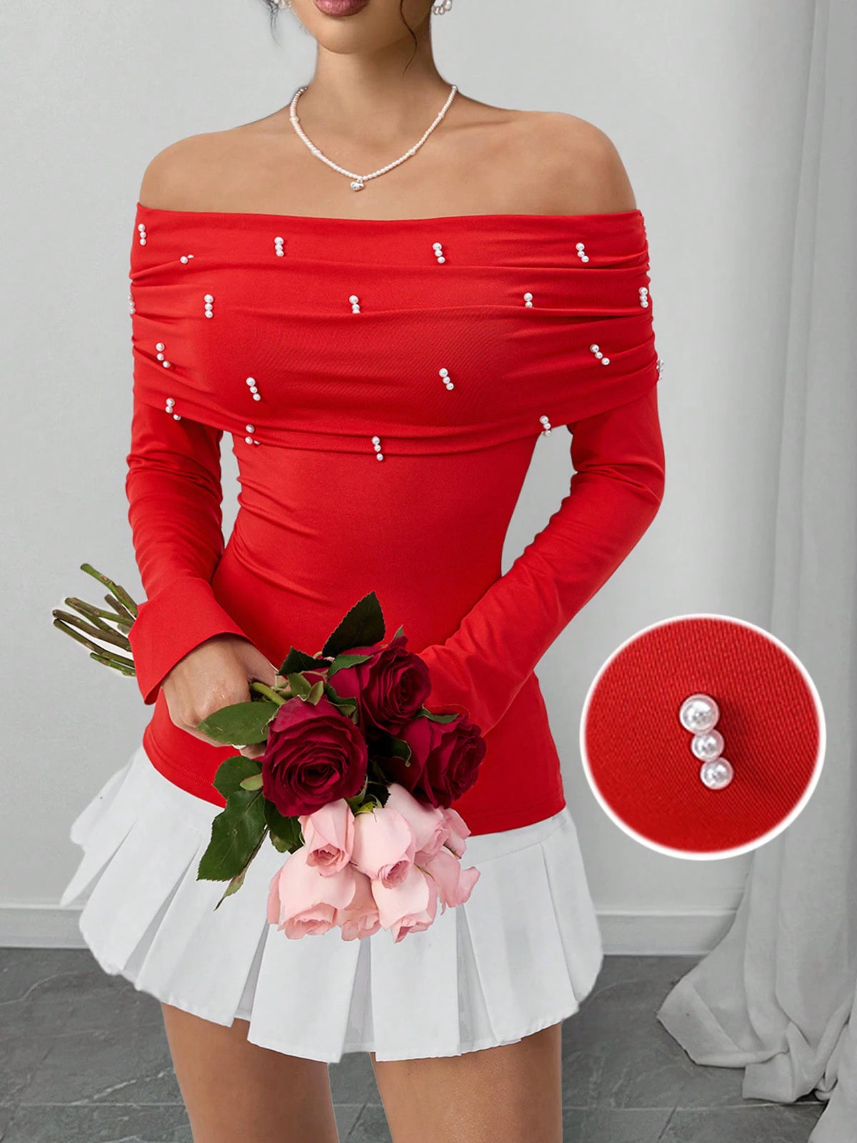 A woman wearing a bright red off-the-shoulder long-sleeve top with small pearl embellishments, paired with a white pleated mini skirt, holding a bouquet of red and pink roses. A circular inset shows a close-up of the pearl embellishment on the red fabric.
