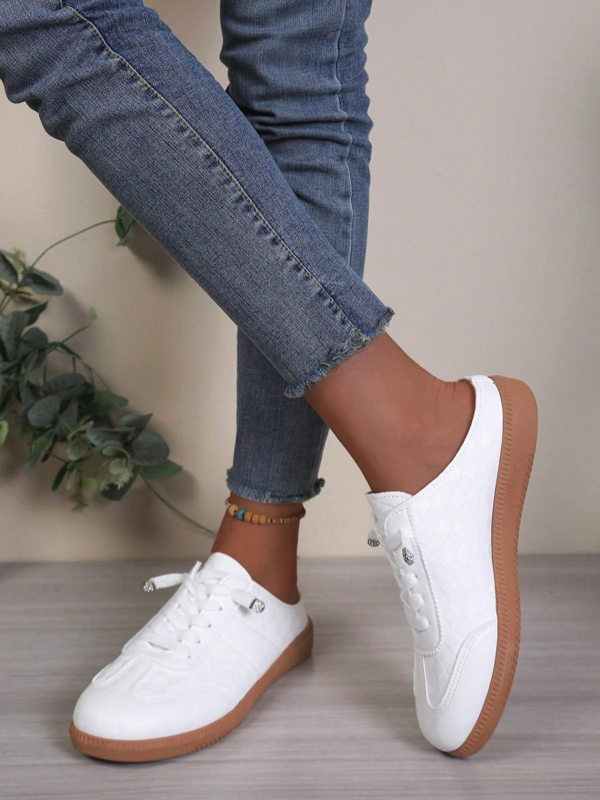 White low-top lace-up sneakers with a gum sole, shown on a model wearing blue jeans against a neutral indoor background.