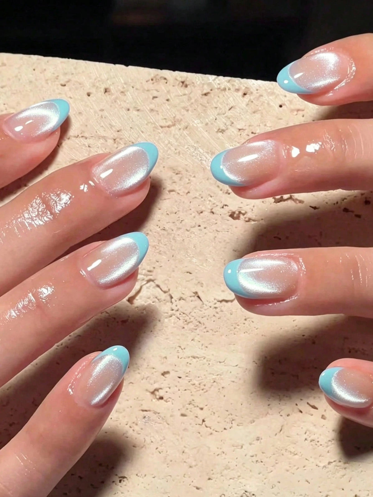 Close-up of hands with manicured nails featuring a glossy white cat-eye finish and a light blue French tip design, resting on a textured beige stone surface.