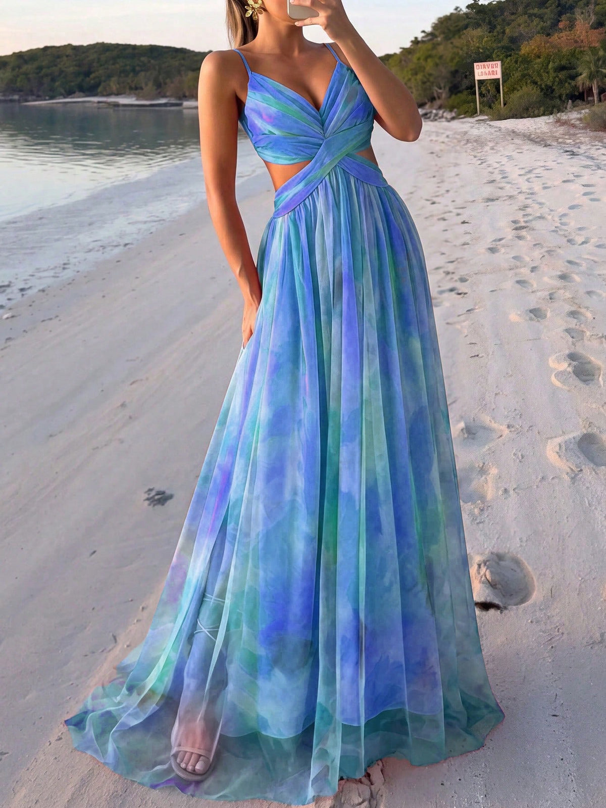 A woman wearing a sleeveless, floor-length A-line dress with a blue and green tie-dye pattern, standing on a sandy beach.