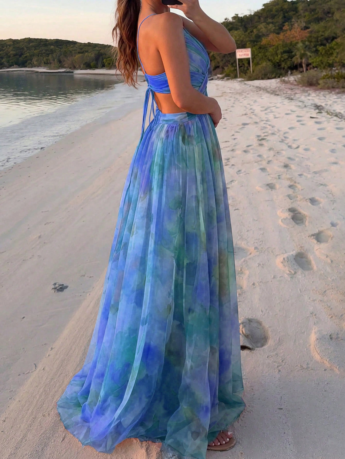 A woman stands on a sandy beach wearing a flowing halter-neck maxi dress with a blue, green, and purple tie-dye watercolor pattern.
