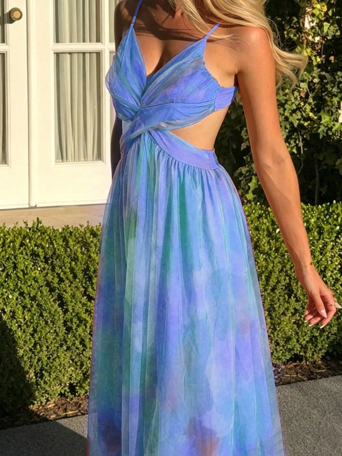 A woman wearing a sleeveless, V-neck maxi dress with a twisted front detail and side cutouts. The dress has a watercolor-like tie-dye pattern blending shades of blue, purple, and teal.