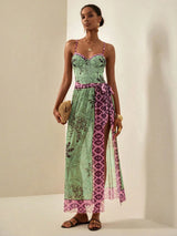 A woman models a sleeveless maxi dress with a light green tropical print and magenta-purple geometric patterned panels, holding a woven clutch against a warm beige studio background.