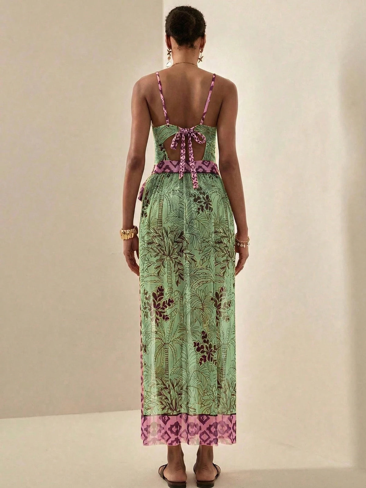 A woman viewed from the back wearing a light green maxi dress with a botanical print, pink patterned straps and trim, and an open back tied with a bow.